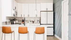 A modern kitchen with white cabinets, a central island with bar stools, a refrigerator, and a hanging light fixture.