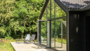 A modern outdoor deck with glass doors leading to a covered area, surrounded by lush greenery. - 5 bedroom nice home in Græsted (Græsted)