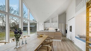A modern kitchen with white cabinetry and a large island, a dining area with a wooden table and chairs, and a view of the outdoors through large windows. - 5 bedroom nice home in Græsted (Græsted)