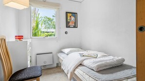 A small, neatly organized bedroom with a single bed, a chair, a radiator, a window with a view of greenery, and a framed picture on the wall.