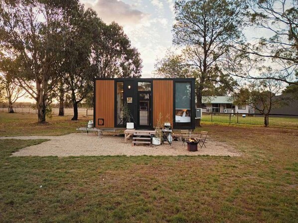 A modern, compact cabin with a wooden exterior, large windows, and a small porch area with outdoor seating. - Ballarat Bliss by Tiny Away (Cardigan)