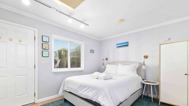 Un dormitorio con cama, mesita de noche, lámpara, ventana con persianas y puerta.