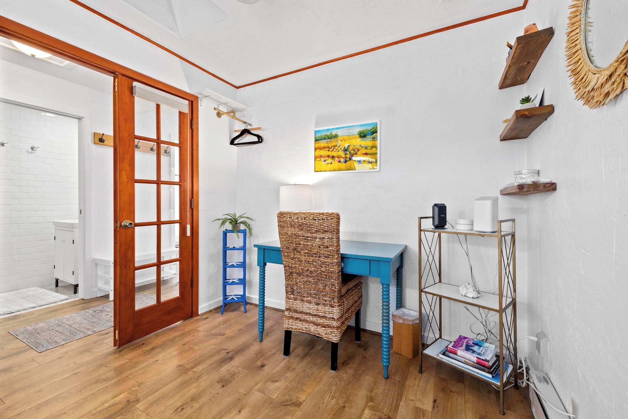 A room with a wooden door, a blue desk, a wicker chair, a white wall with a painting, and a shelf with books and a lamp.