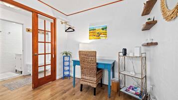 A room with a wooden door, a blue desk, a wicker chair, a white wall with a painting, and a shelf with books and a lamp.