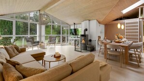 Een ruime woonkamer met een grote bank, eettafel en stoelen, een open haard en grote ramen met uitzicht op de omgeving.