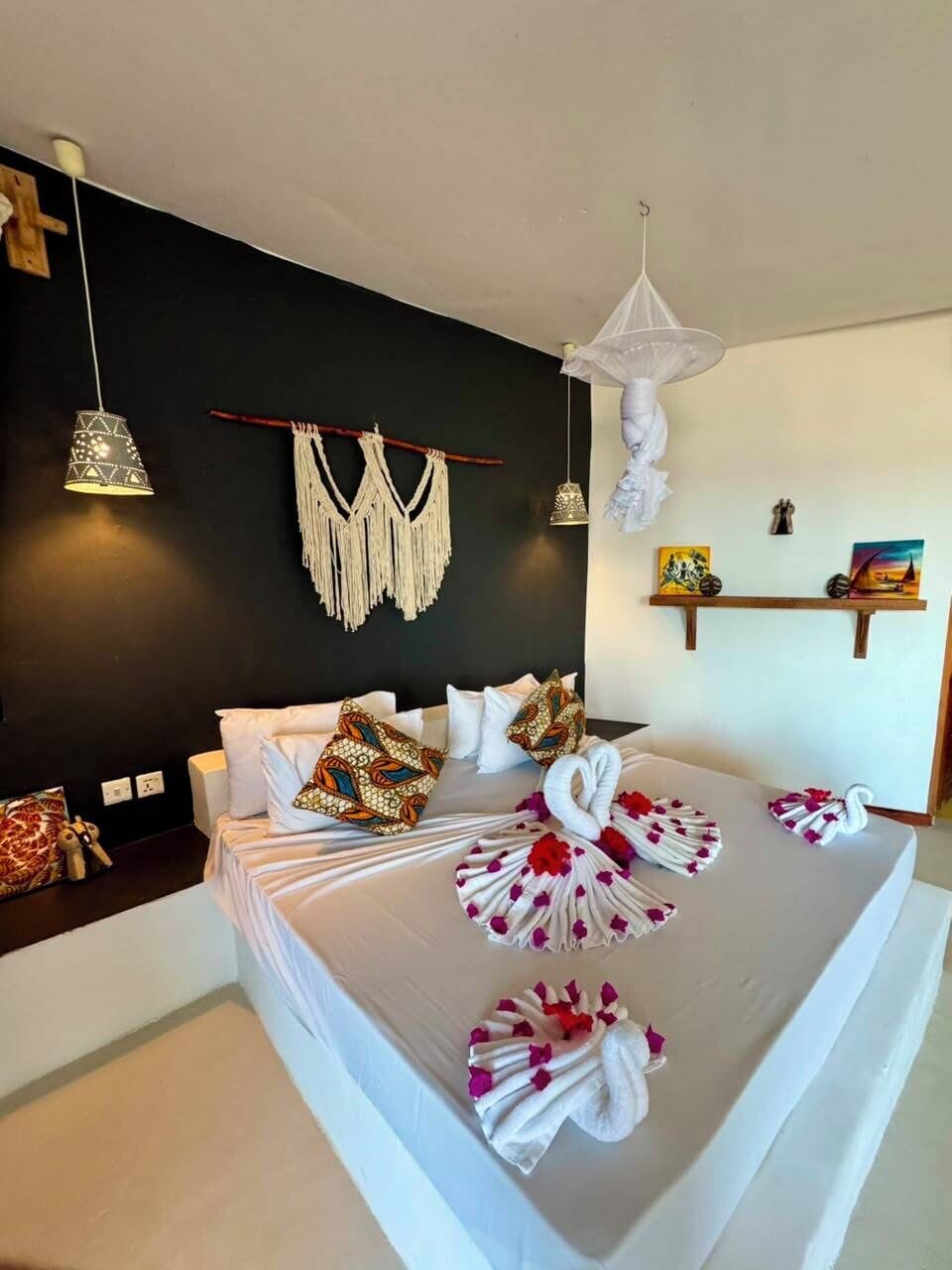 Un dormitorio moderno con cama, un tapiz de macramé, luces colgantes y una estantería con objetos decorativos.
