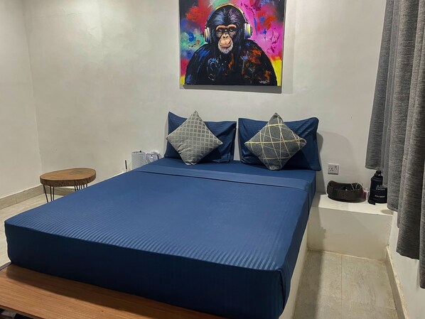 A bedroom with a large bed, two pillows, a small table, a painting on the wall, and a curtain.