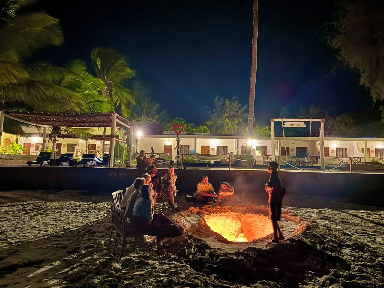 A group of people gathered around a fire pit on a beach at night, with a building in the background.