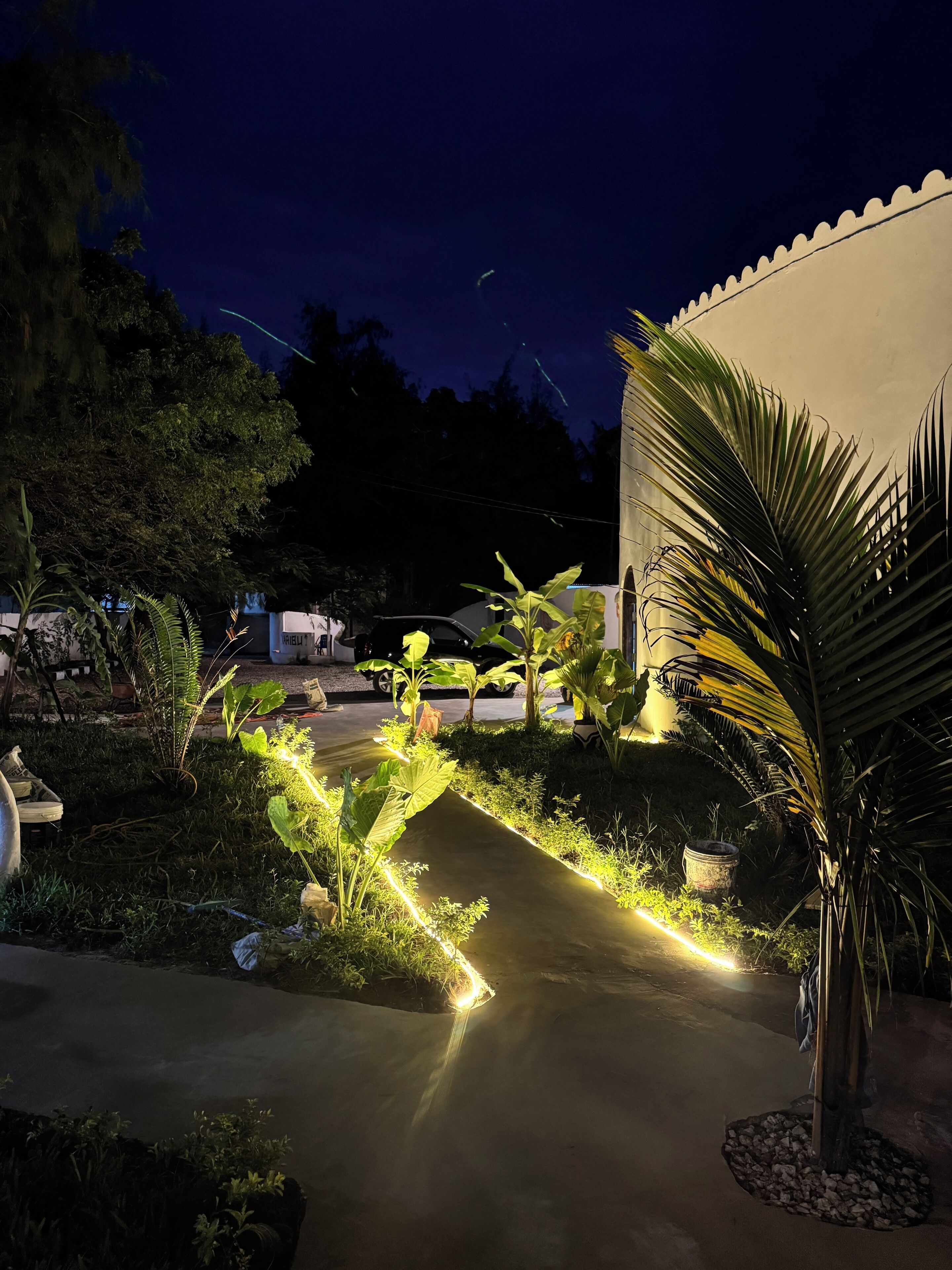 A nighttime outdoor area with a pathway lit by linear lights, surrounded by greenery and a building with a white facade.