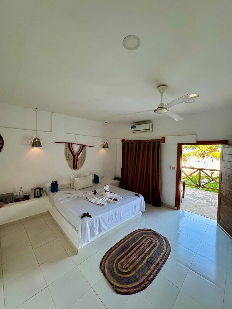 A bedroom with a bed, a ceiling fan, an air conditioning unit, a window with curtains, and a view of the outdoors.