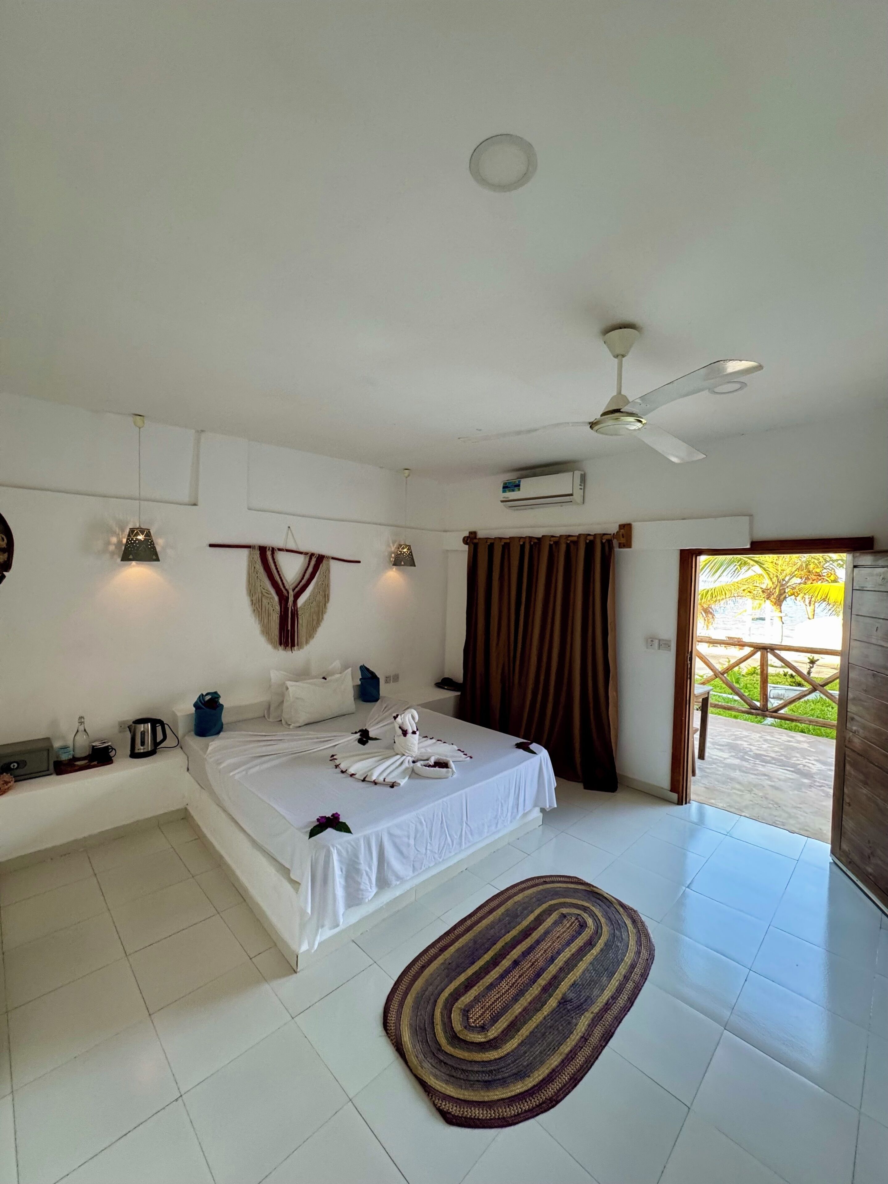 A bedroom with a bed, a ceiling fan, an air conditioning unit, a window with curtains, and a view of the outdoors.