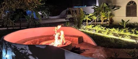 A nighttime outdoor area with a fire feature, a white building with a rounded corner, and surrounding greenery.