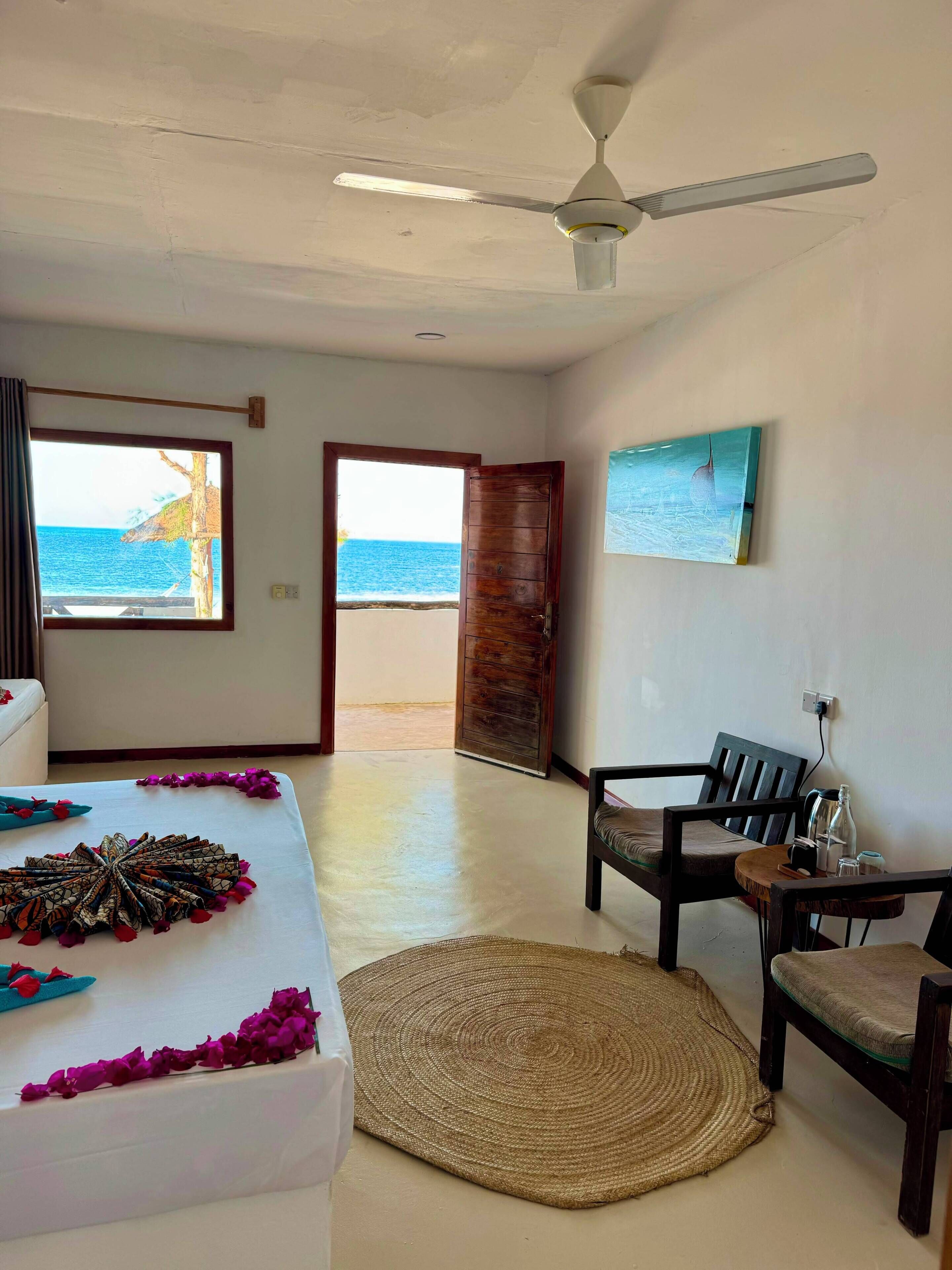 A room with a bed, a ceiling fan, a view of the sea through a door, a chair, and a table with items on it.
