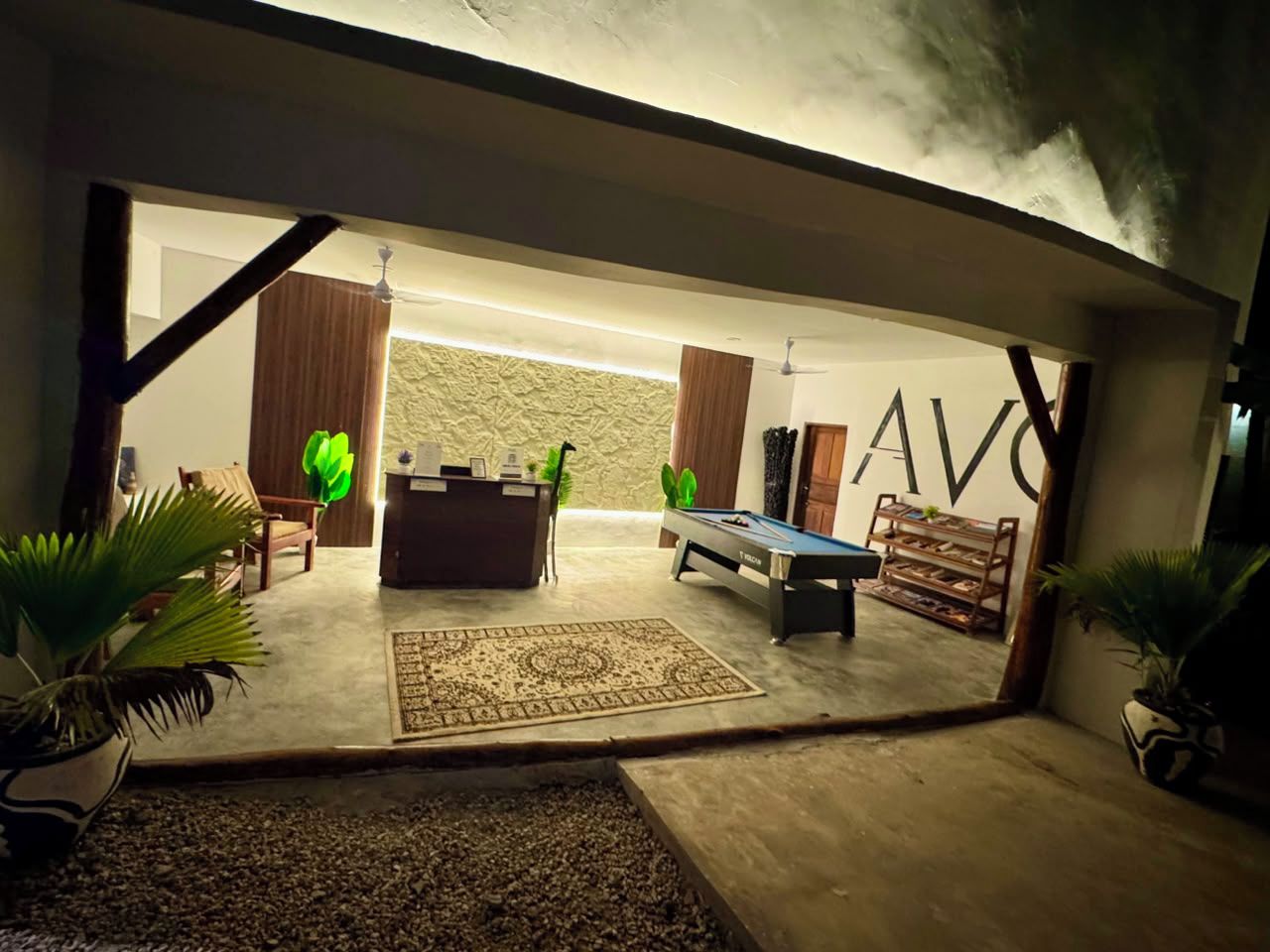Habitación con mesa de billar, sofá y una silla. Se observa un ventanal con cortinas y una pared donde se aprecian las letras 'AV'.