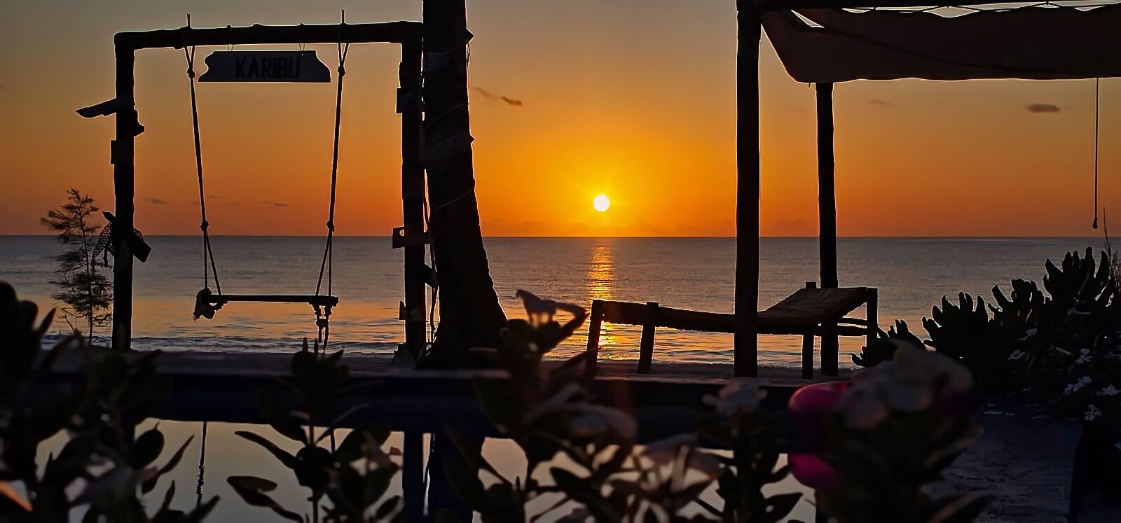 A beach scene with a swing set and a chair facing the sunset over the ocean.