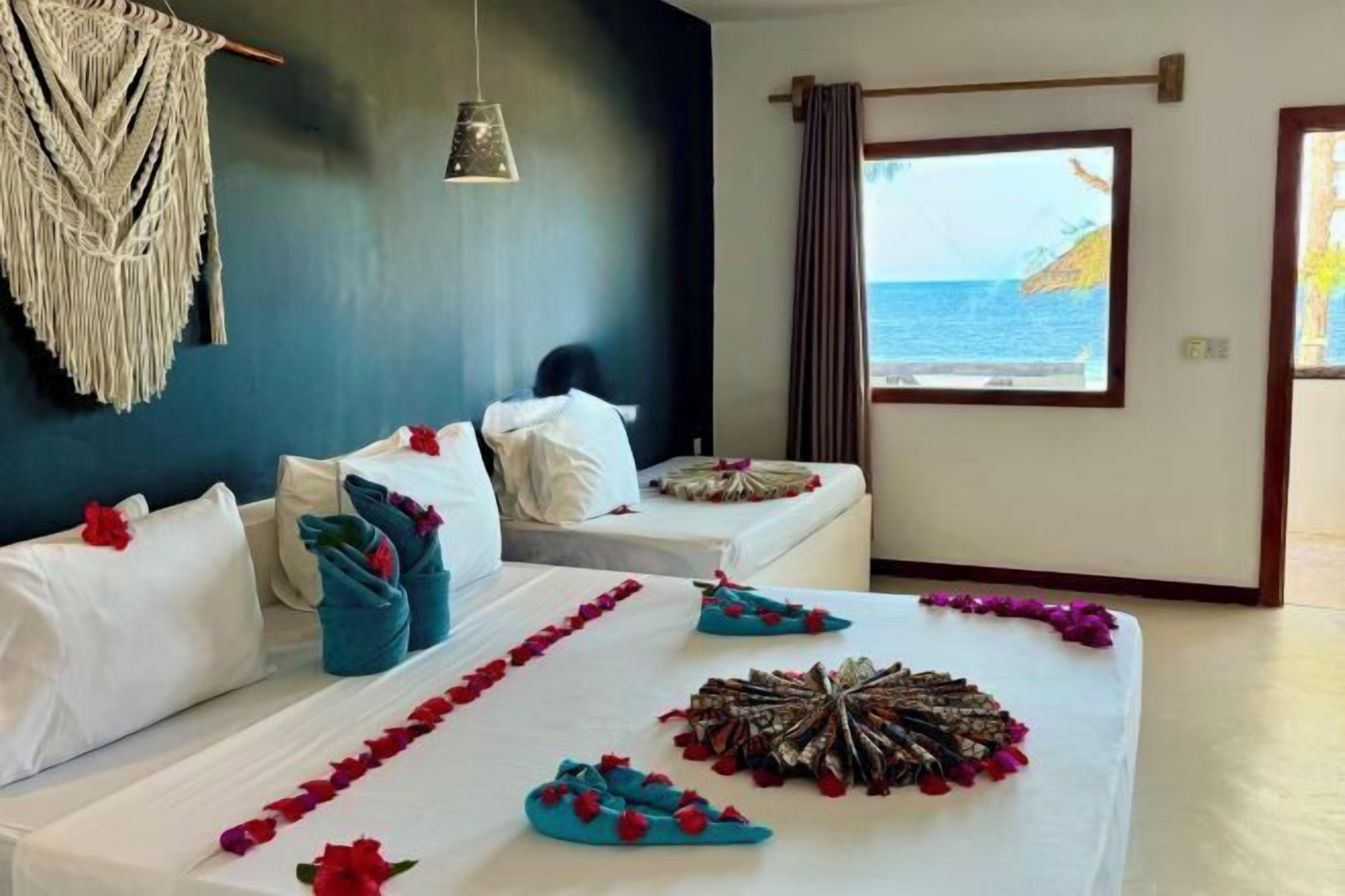 A hotel room with two beds, a view of the sea, a macrame wall hanging, and a hanging light fixture.