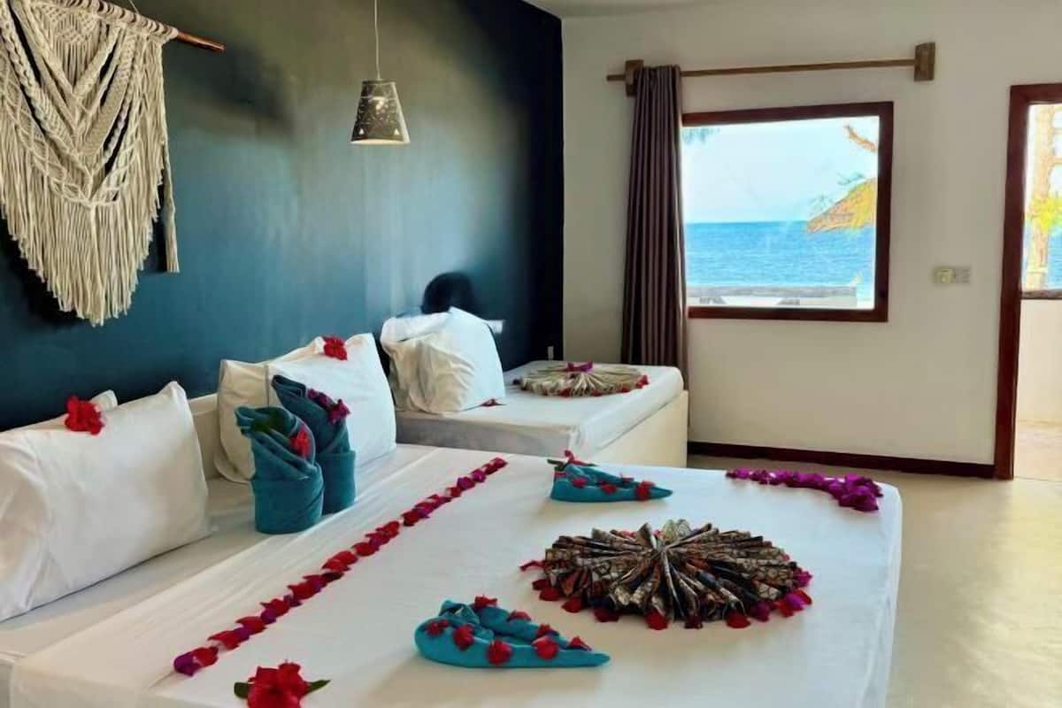 A hotel room with two beds, a view of the sea, a macrame wall hanging, and a hanging light fixture.