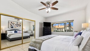 A bedroom with a large bed, a ceiling fan, a dresser, a TV, and a window with a view of houses. - Near Coachella Two Bedroom Two Bath Condo, Steps from the Pool & Spa, Desert Falls CC (Palm Desert)