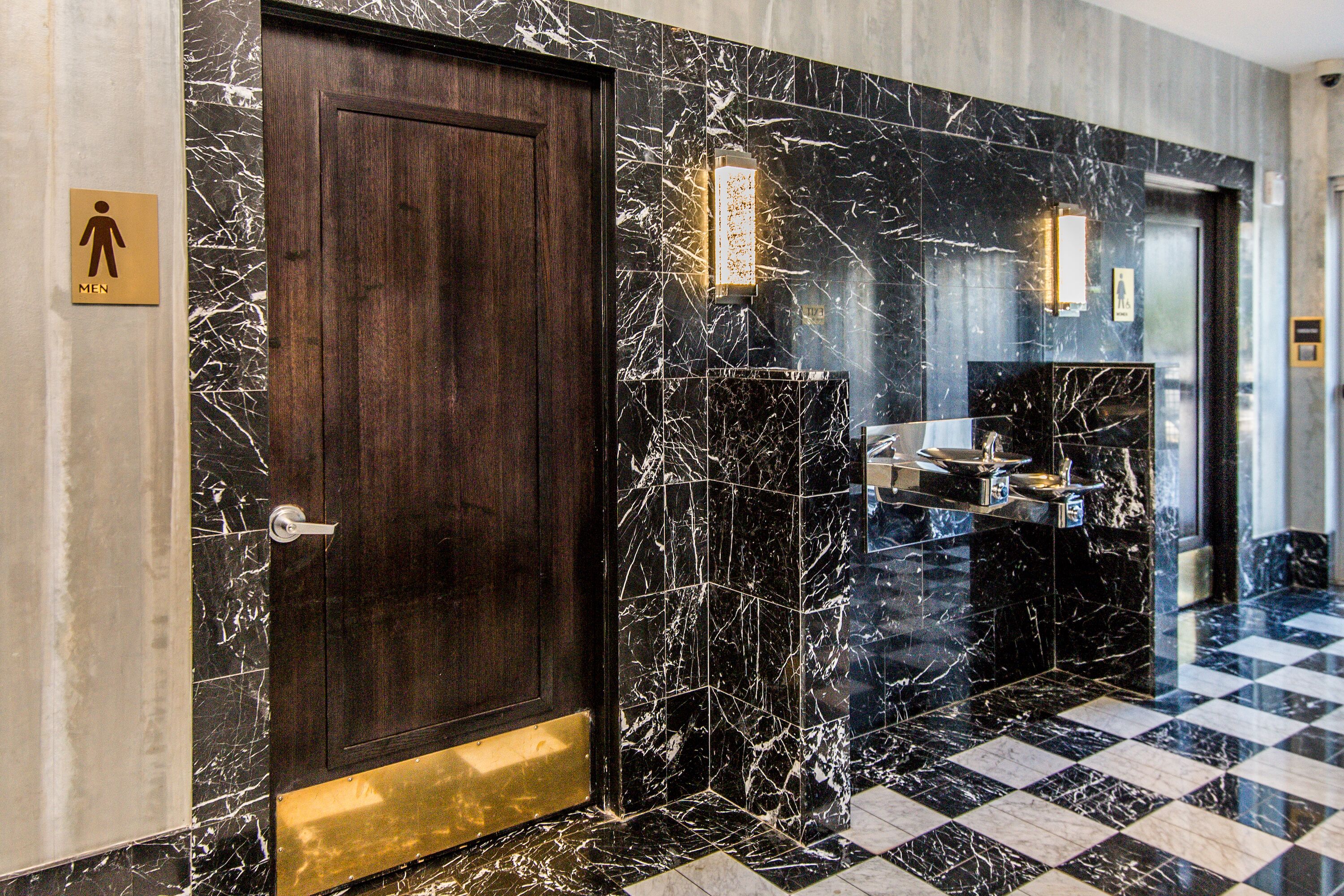 A hotel lobby with a checkered floor, a marble wall with a door, and a men's restroom sign.