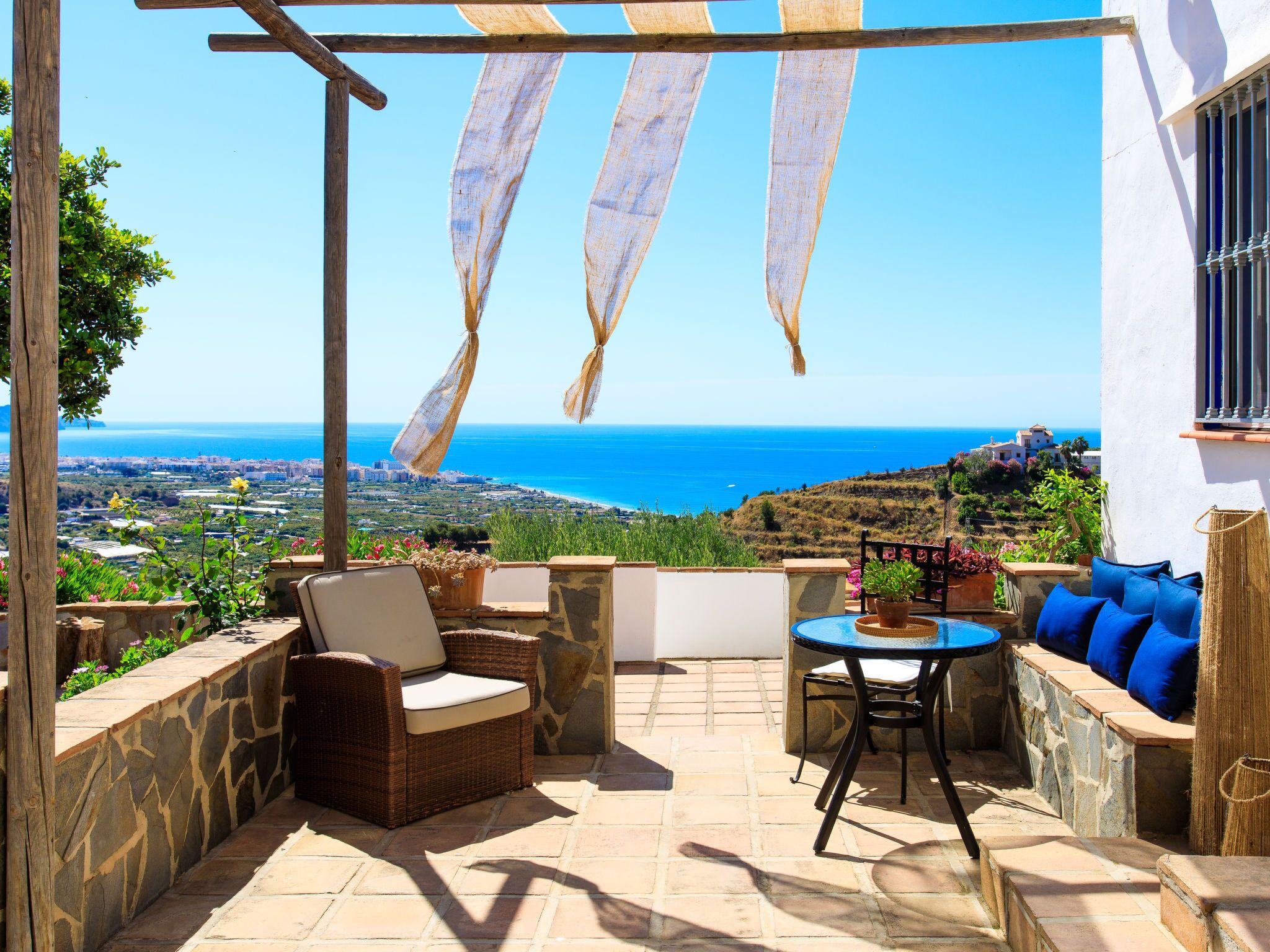 A patio with wicker furniture, a round table, and a view of the sea and landscape.