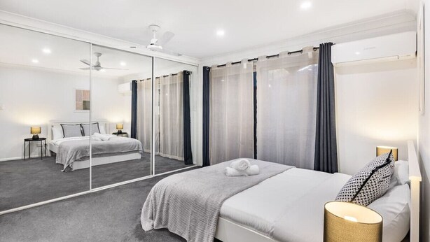 Ein modernes Hotelzimmer mit einem großen Spiegel, einem Bett mit grauer Decke, einem Nachttisch mit Lampe und einem Deckenventilator.