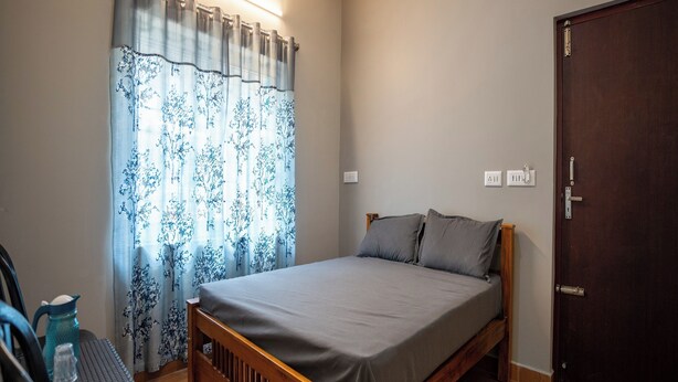 A bedroom with a wooden bed, grey bedding, a window with floral curtains, a bedside table with a blue water bottle, and a dark wooden door. - Hill Lock Mist (Kodaikanal)