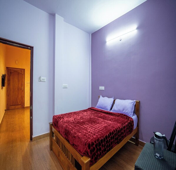 Un dormitorio con cama de madera, colcha estampada roja, pared morada, lámpara de techo blanca y puerta de madera.