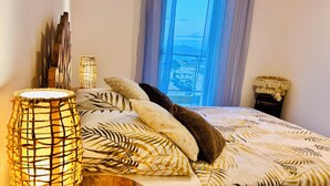 Un lit recouvert d’une couvre-lit à motifs de feuilles, une lampe décorative et une vue sur le paysage urbain par la fenêtre.