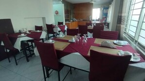 Un comedor con mesas dispuestas para cuatro, sillas rojas y una cocina al fondo.
