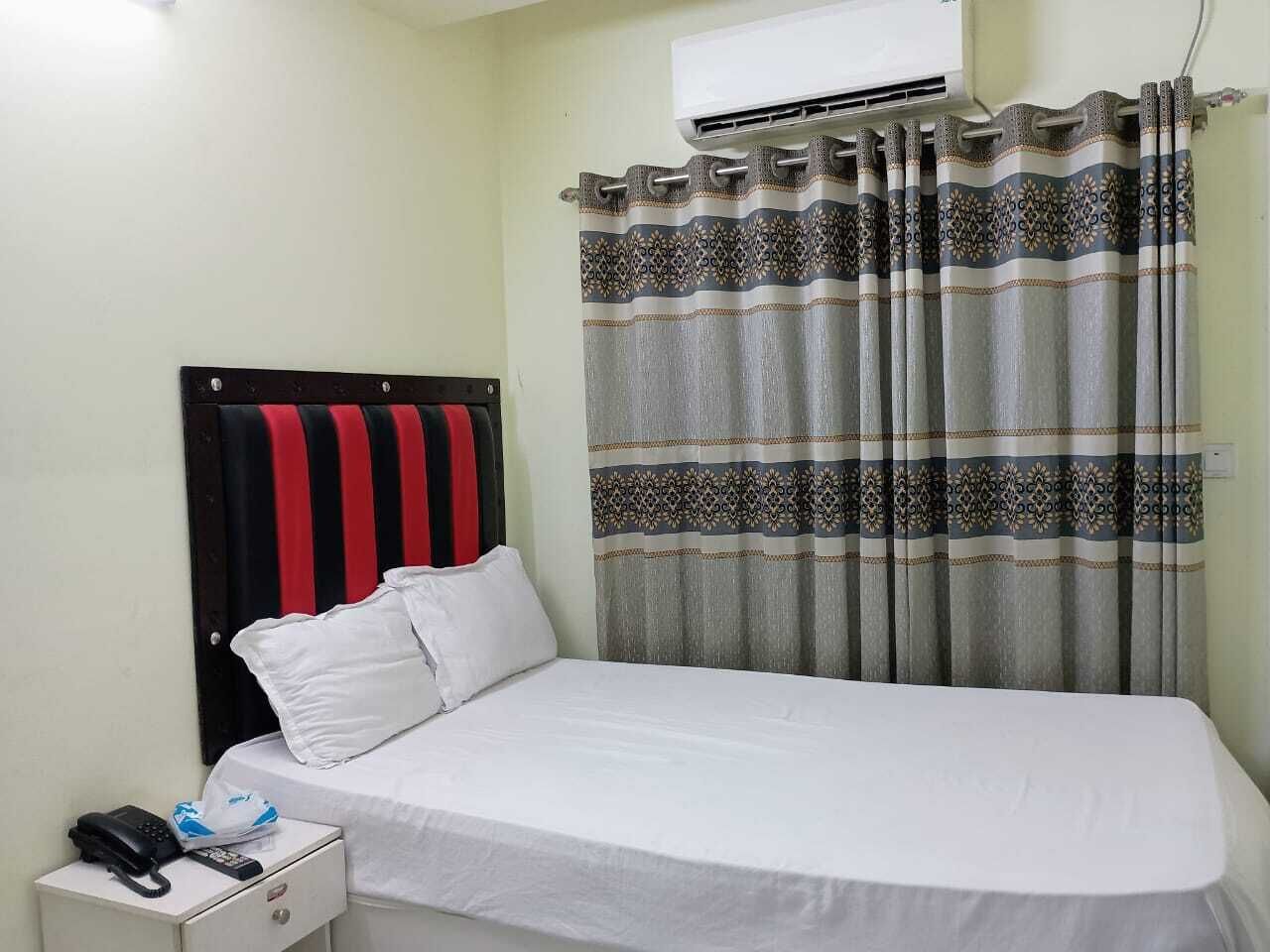 A single bed with a white mattress and pillows, a bedside table with a phone and a tissue box, and a patterned curtain.