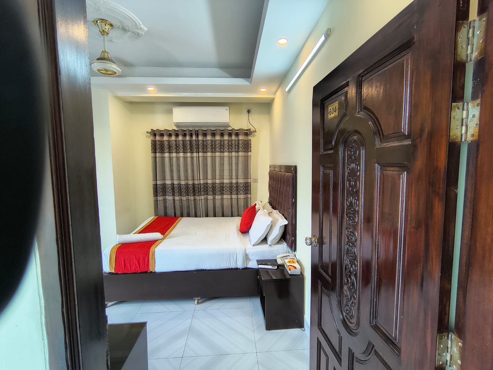 A hotel room with a bed, an air conditioning unit, a bedside table, and a door with a number plate.