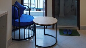 Una habitación pequeña con una mesa redonda, una silla azul, un balcón con vistas a edificios y un baño con lavabo y espejo.