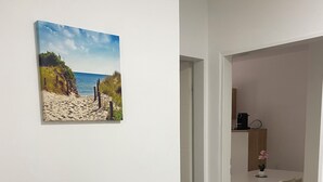 Une pièce avec un plancher en bois, une table blanche avec des chaises, une petite plante en pot et un tableau représentant une scène de plage accroché au mur.