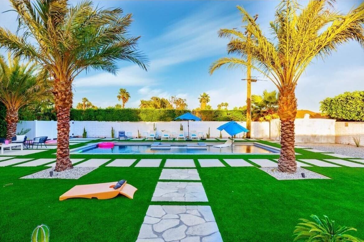 Ein Garten mit Swimmingpool, Palmen und einer Lounge-Ecke mit Stühlen und Sonnenschirmen.