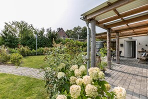 A covered patio area with a brick floor, a wooden pergola, and a view of a house in the background.