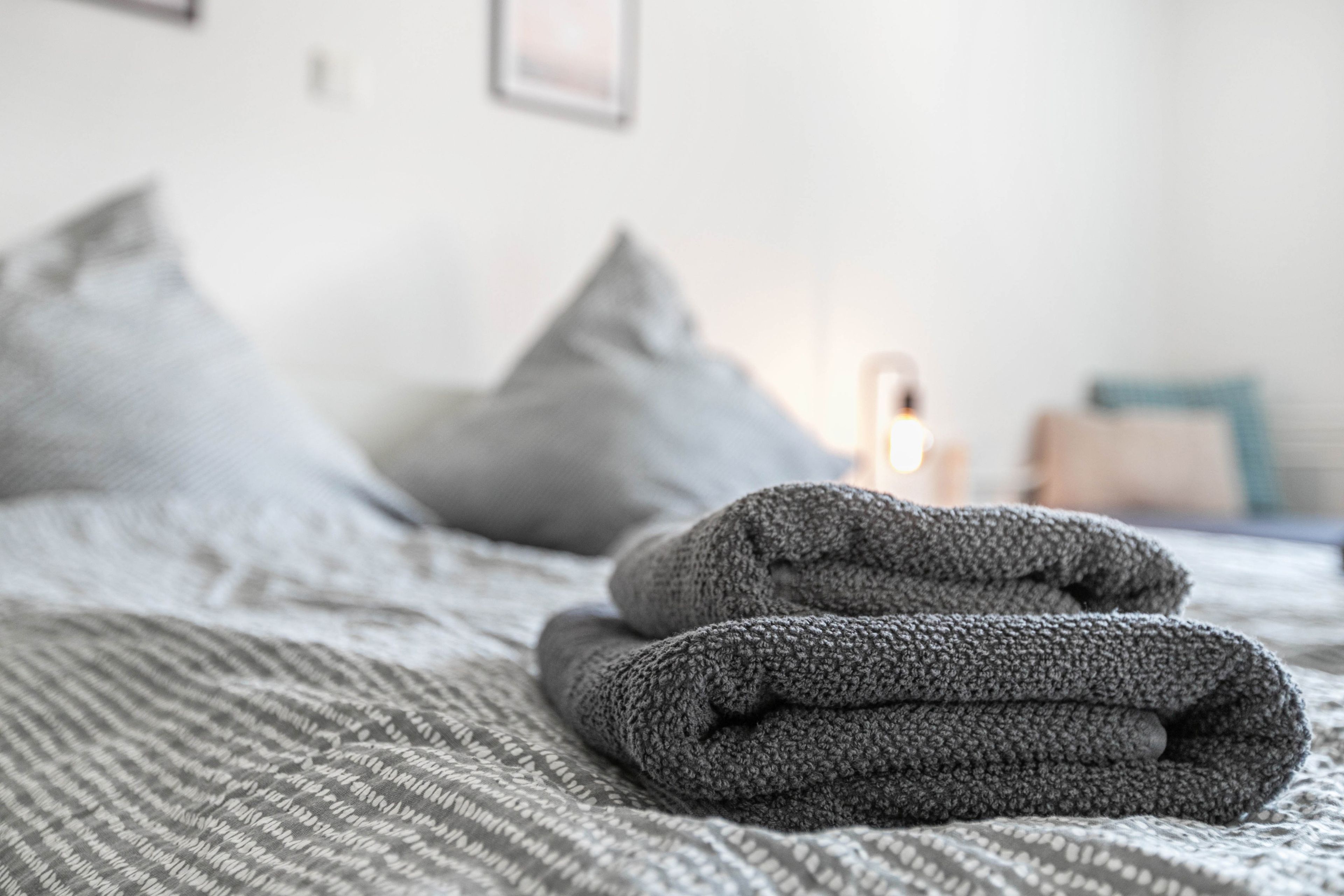 Ein ordentlich bezogenes Bett mit grauer Bettwäsche und Kissen, eine gefaltete graue Decke sowie ein Nachttisch mit einer Lampe.