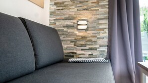 A grey sofa with a patterned pillow, a stone wall with a light fixture, and a grey curtain. - Apartment 'Ferienanlage Kappeln-Kopperby' with Private Terrace, Private Garden and Wi-Fi (Kappeln Kopperby)