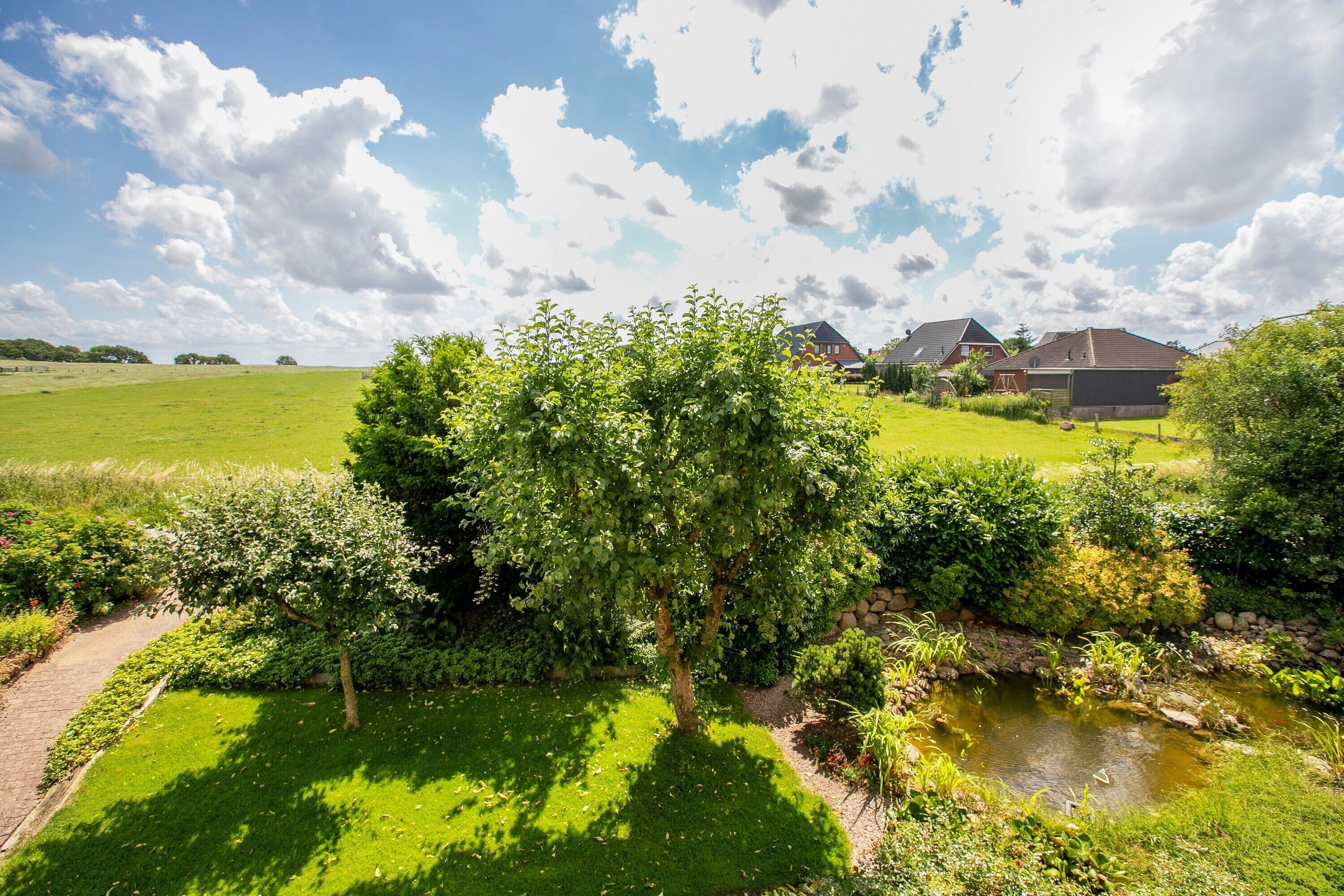 Ländliche Landschaft mit Teich, üppiger Vegetation und Häusern im Hintergrund.