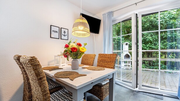 Ein Essbereich mit Korbstühlen, einem weißen Tisch, einer Vase mit Blumen, einem an der Wand befestigten Fernseher und einer Glastür zu einem Garten.