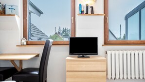 A room with a TV on a wooden cabinet, a chair, a desk, and a window with a view of buildings and trees. - Apartment 'Ferienwohnung Relax' with Private Terrace, Private Garden and Wi-Fi (Stralsund)