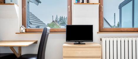 Ein Zimmer mit Fernseher auf einer Holzeinrichtung, einem Sessel, einem Schreibtisch und einem Fenster mit Blick auf Gebäude und Bäume.