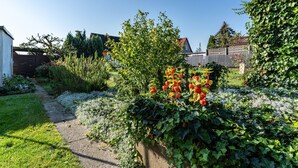 Um jardim com um caminho, um arbusto com flores vermelhas e uma casa ao fundo.