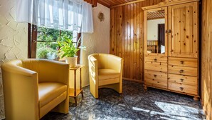 Quarto com paredes e teto de madeira, um guarda-roupa espaçoso, duas poltronas amarelas, uma mesinha e uma janela com uma planta lá fora.