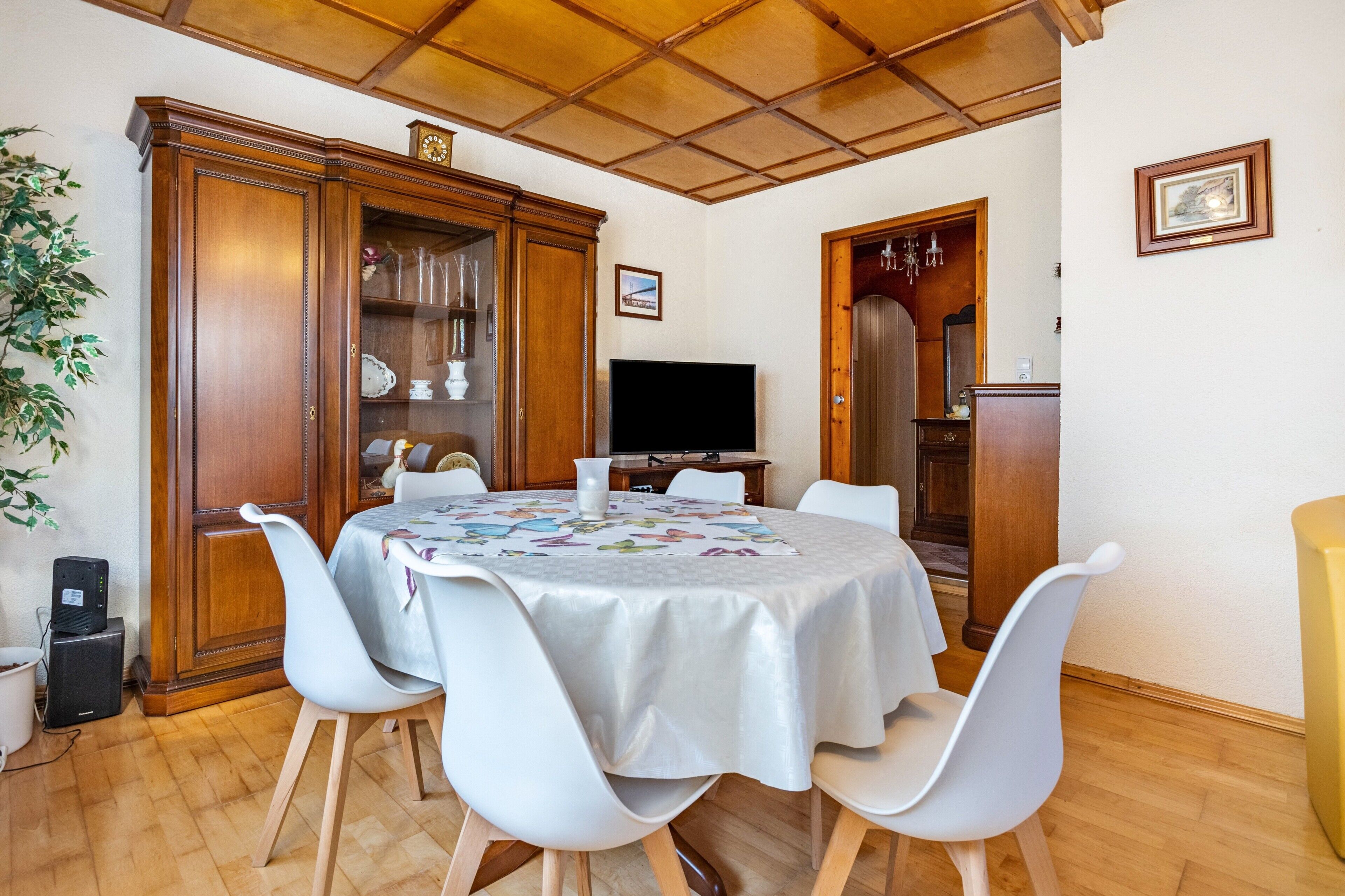 Un comedor con una mesa redonda, sillas blancas, un mueble de madera con cristalería, un televisor y una planta.