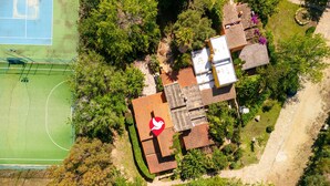An aerial view of a property with a tennis court, a house with a red roof, and surrounding greenery.