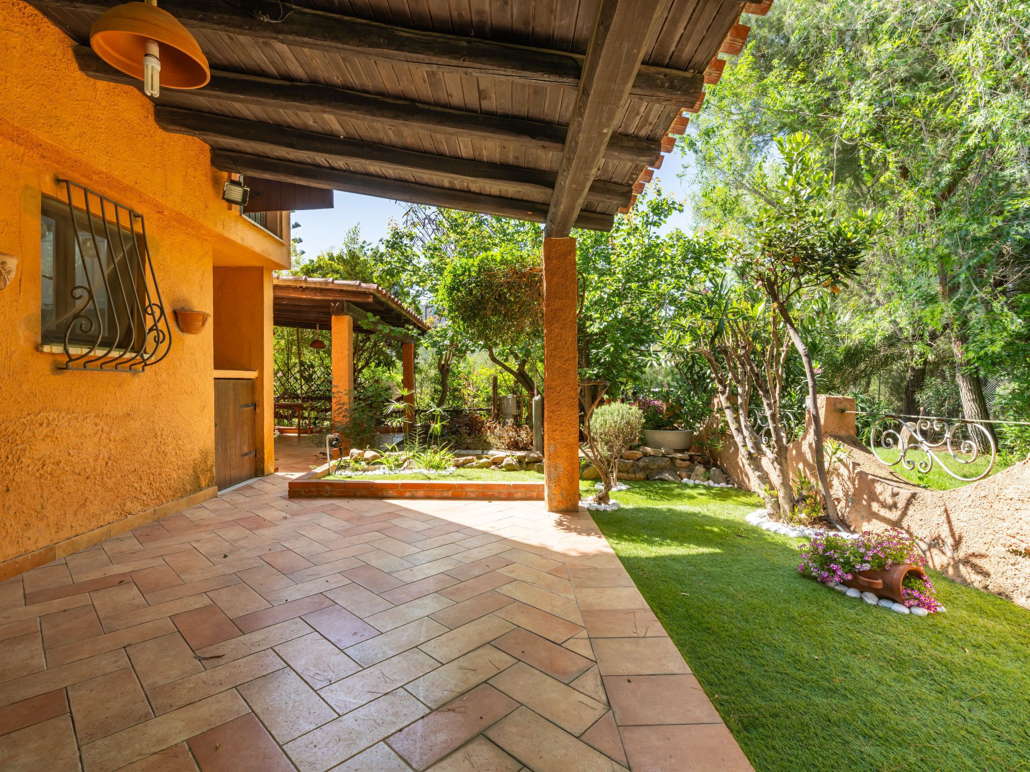 Un patio couvert doté d’un sol carrelé, d’une pergola et d’un jardin agrémenté de diverses plantes et d’un petit arbre.