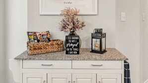 Una encimera de cocina con una ilustración enmarcada de una casa, una cesta de revistas, un jarrón con flores secas y un letrero con una cita.