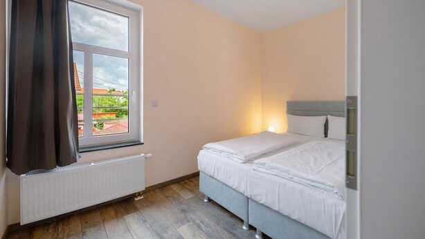 Quarto de hotel com uma cama de solteiro, janela com cortinas, radiador e luminária fixada na parede.