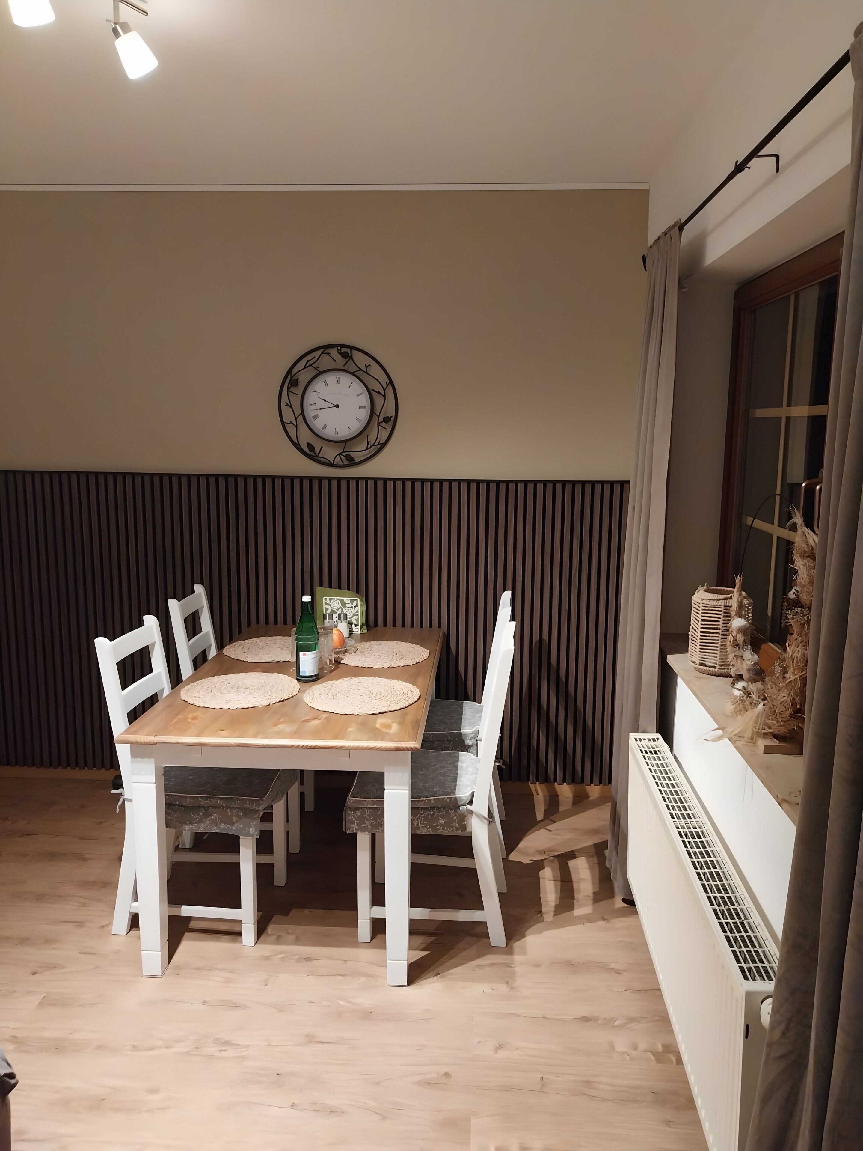 Een eethoek met een houten tafel, witte stoelen, een wandklok en een raam met gordijnen.