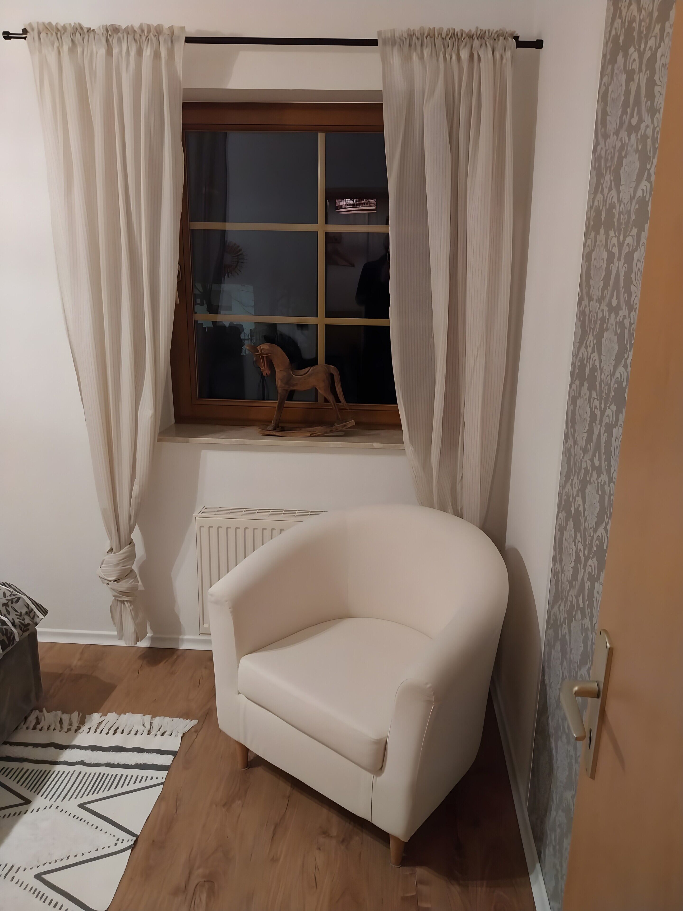 A corner room with a white armchair, a radiator, a window with curtains, and a patterned rug.